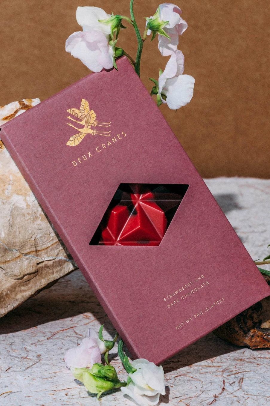 Dark chocolate and strawberries bar  surrounded by flowers and rocks on a textured surface ©Deux Cranes