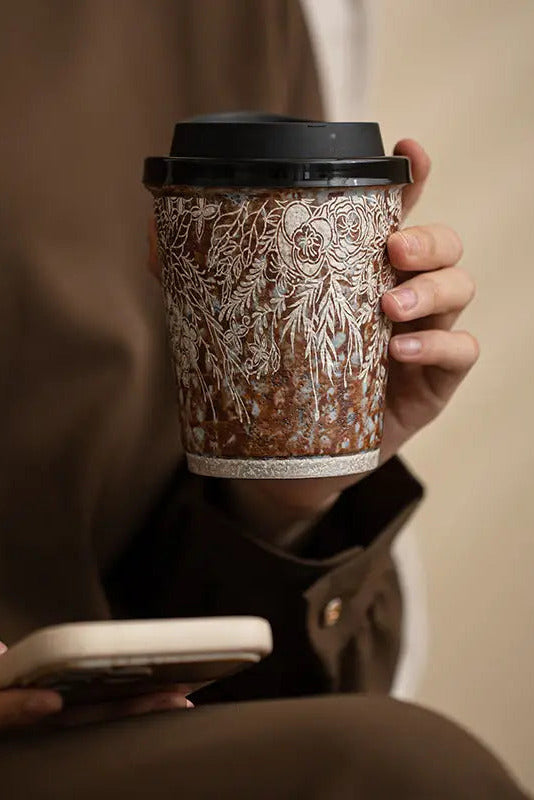 A person in a brown blazer holds a ceramic coffee cup with a black lid and a white floral pattern, while looking at a smartphone. ©Haku