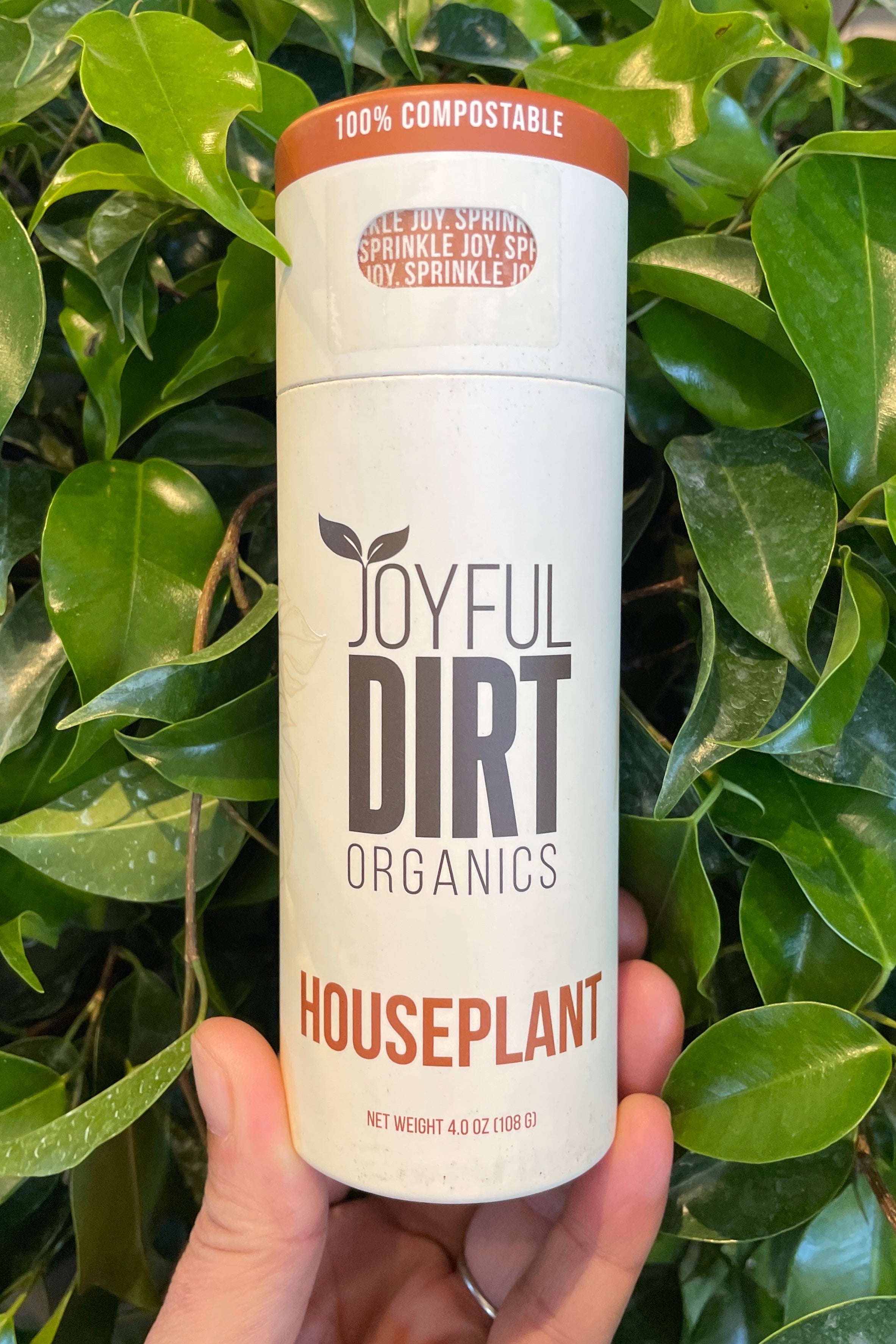 Joyful dirt houseplant fertilizer being held in front of ficus leaves. 