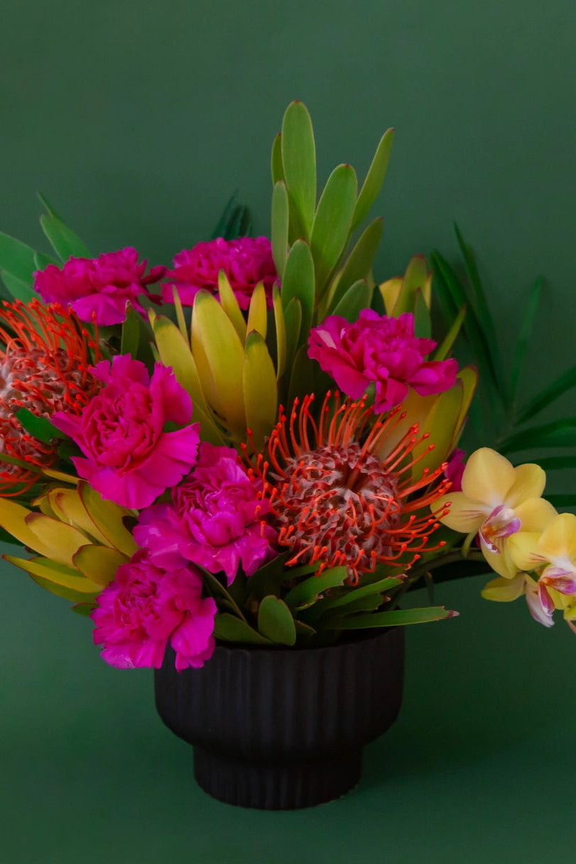 Bouquet of colorful flowers including pink, red, and yellow in a Presley Pedestal Planter black vase on a green background ©Jungle Club