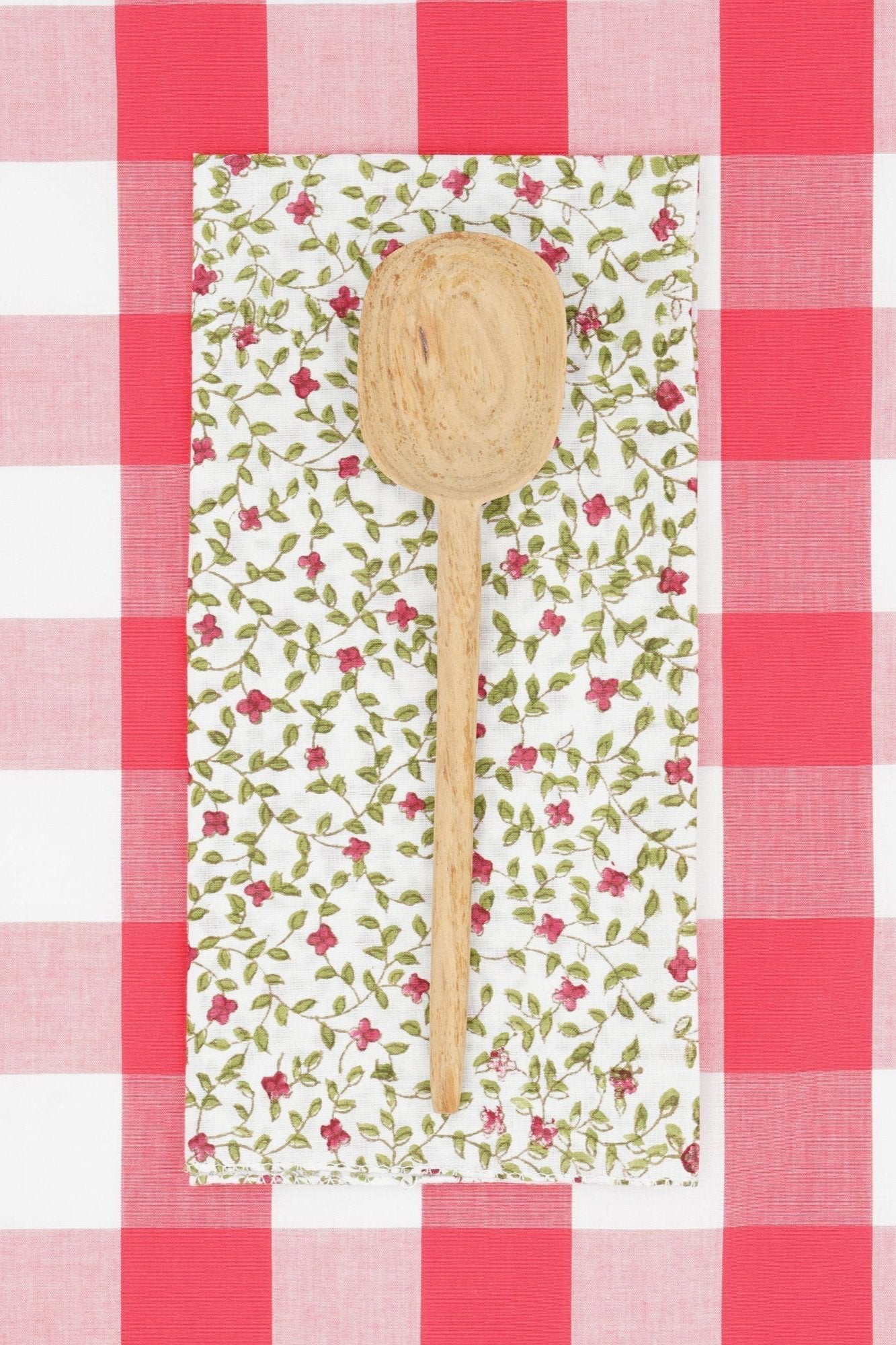 Suzette napkin Alchemille Peonie folded into a rectangle, wood spoon on top and checkered red and white print behind.