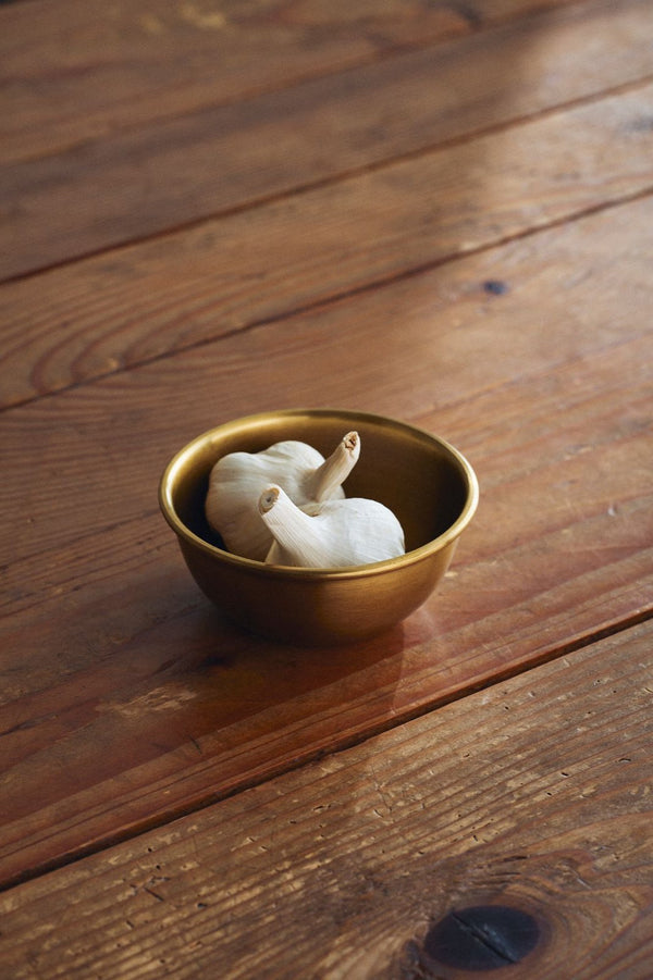 The Small Brass Bowl by Fog Linen Work with two garlic heads inside sitting on a wood slatted table. 