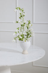 A white Magnolia vase with white bell flowers inside on a white table and white walls.