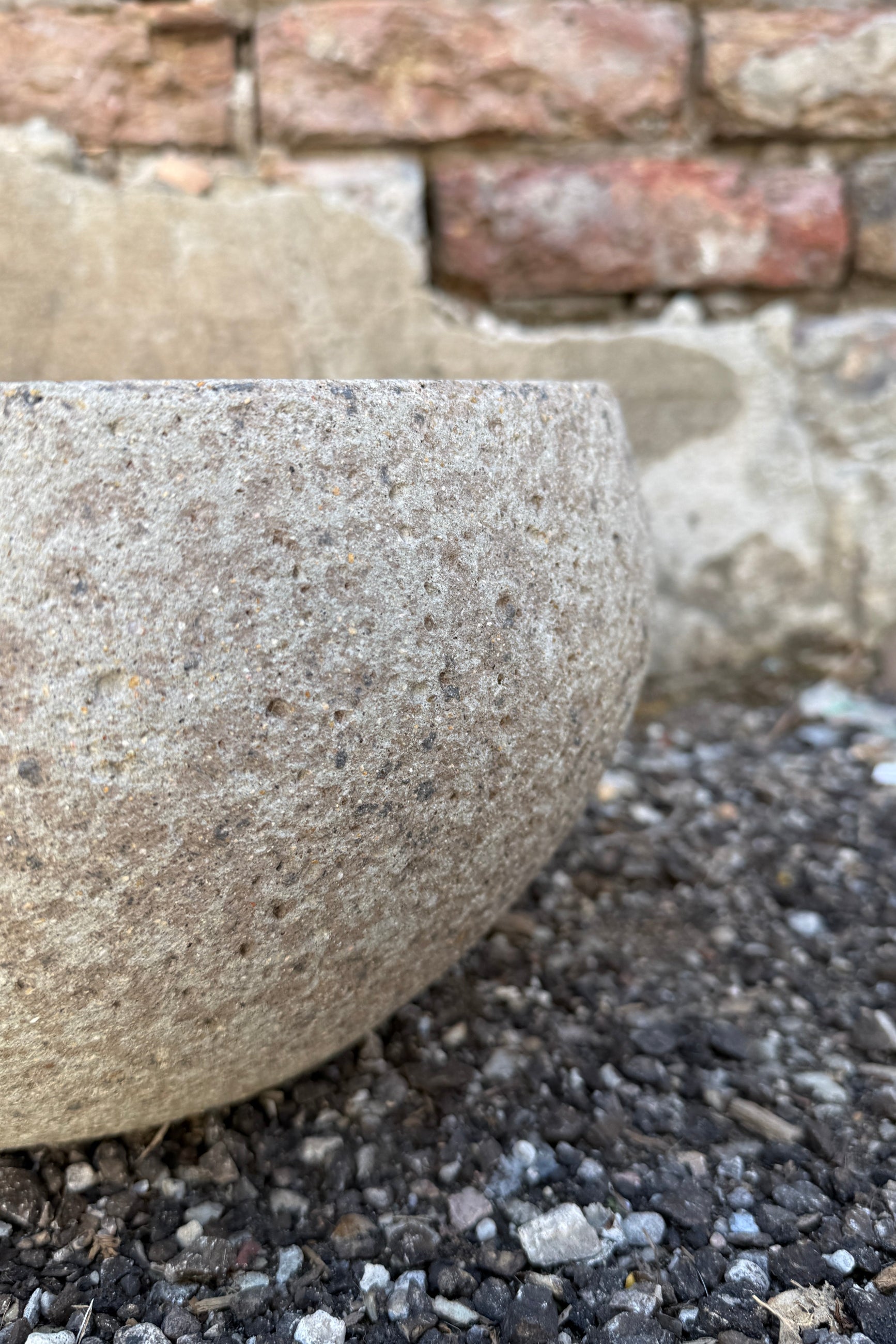 Close-up of a textured Low Ficonstone Greige Bowl Small on a gravel surface with a brick wall in the background. ©Sprout Home