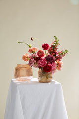 Two Ravish vases on a white cloth covered pedestal with the vase in front filled with various red to pink flowers. 