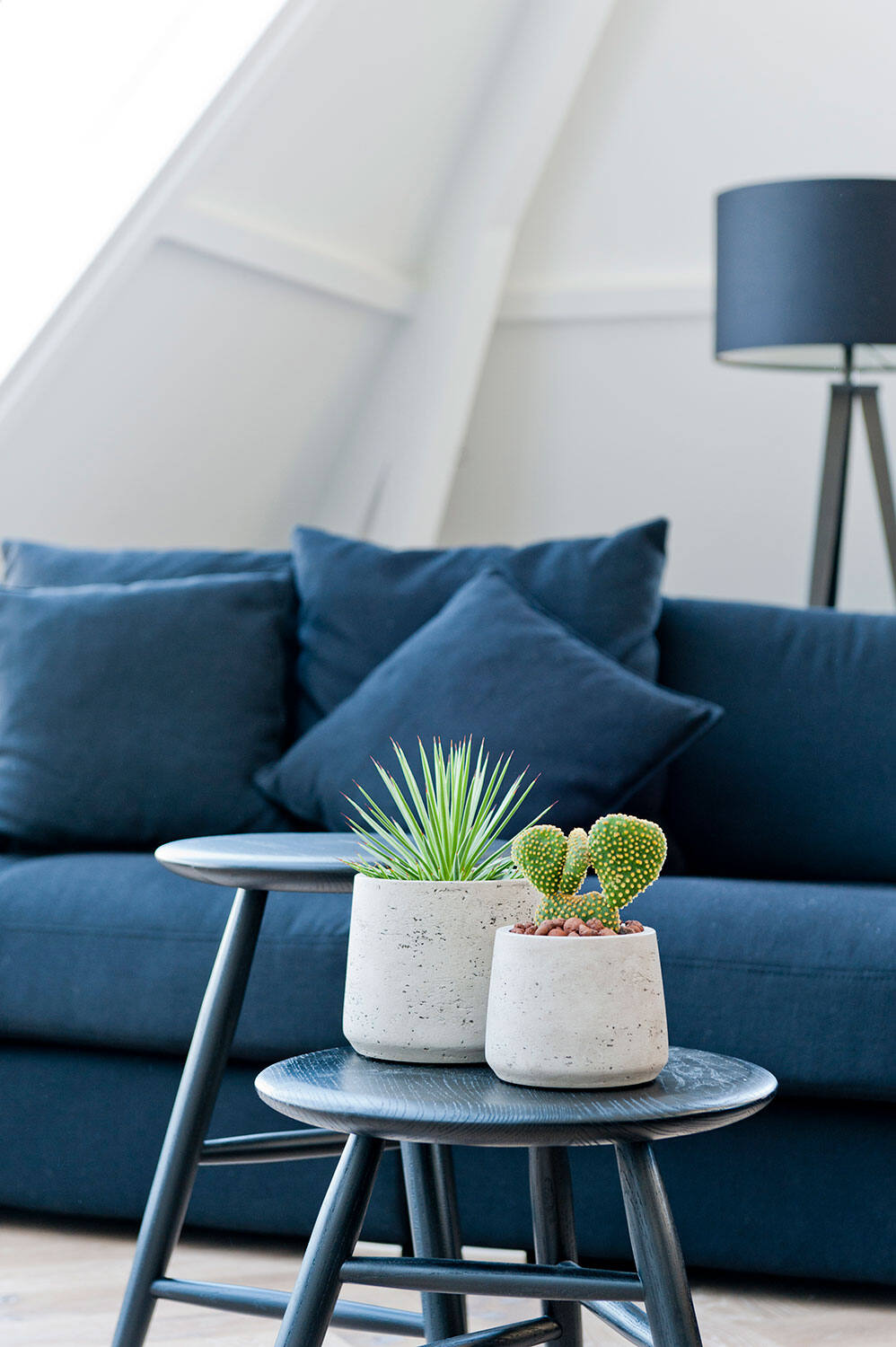 Two different sized Patt Pots in Grey washed both potted with succulents and cactus on a wooden stool in front of a blue sofa.  By Pottery Pots