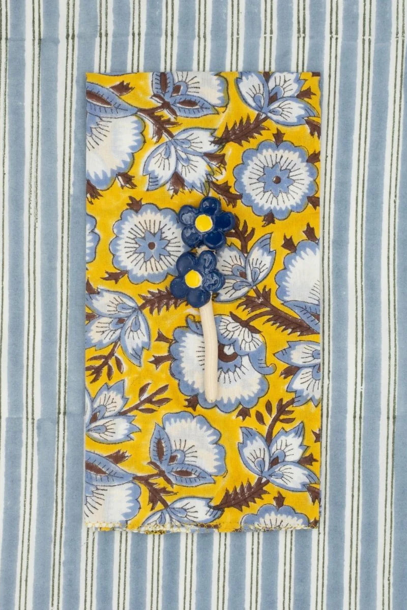 Cotton Napkins Delphinium Moutarde  sitting on a blue striped cloth with a flower on top ©Suzette a Table