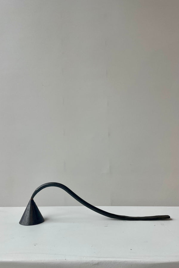 Photo of a black metal candle snuffer on a white surface against a white wall. The handle curves up and then down to rest on the same surface.