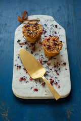 The brass cake server by Fog Linen Work sitting on a serving board with two muffins.