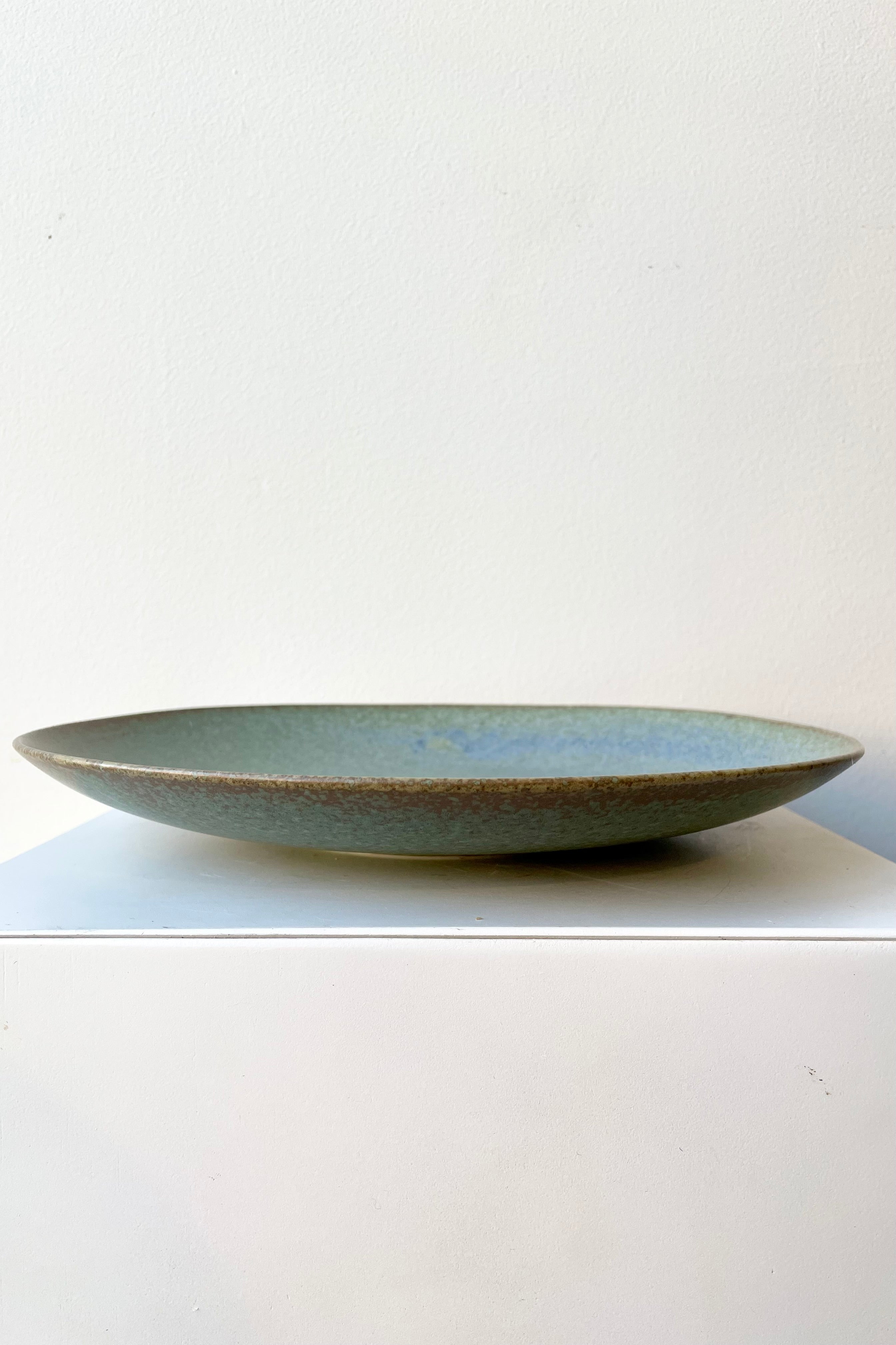 The Shiho plate in terra green against a white wall on a white pedestal. 