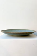 The Shiho plate in terra green against a white wall on a white pedestal. 