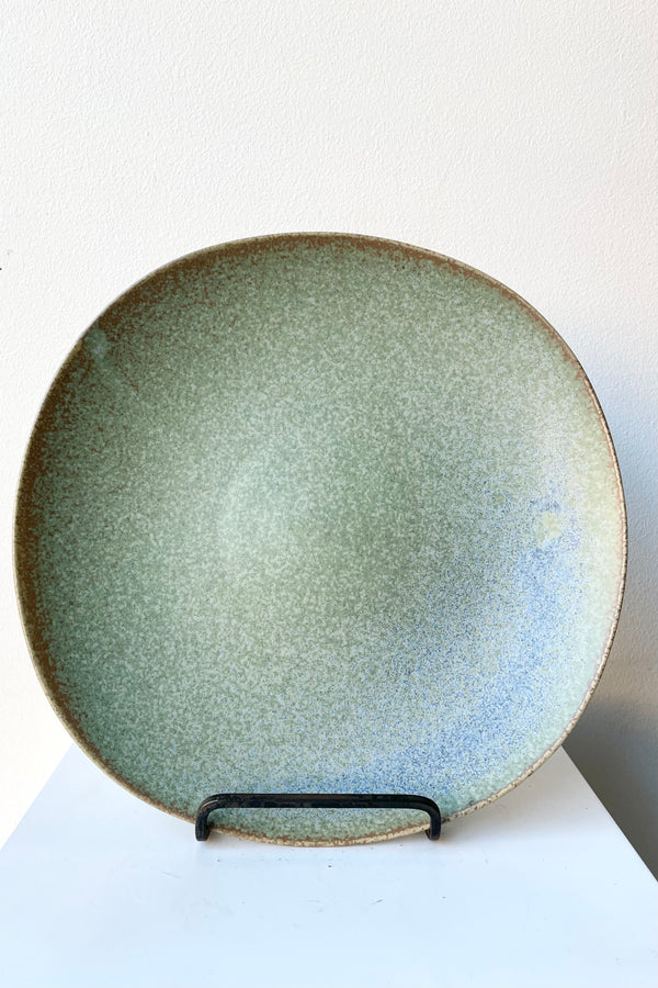 The Shiho plate in terra green against a white wall on a plate stand.