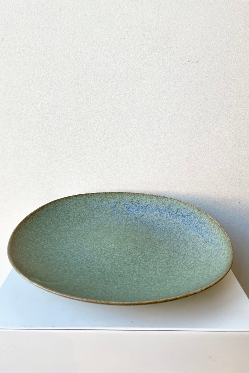 The Shiho plate in terra green against a white wall sitting on a white pedestal.