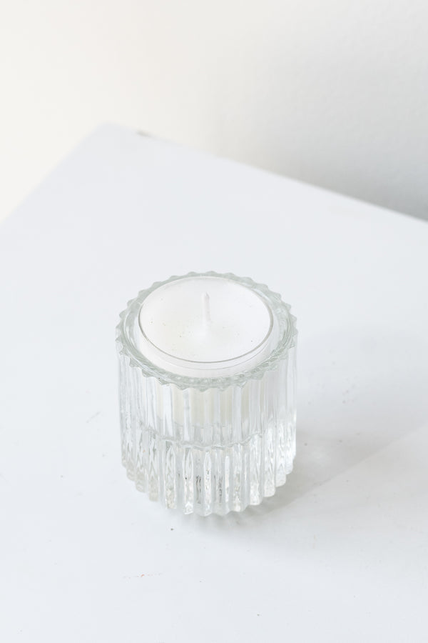 White tea light candle inside clear pleated glass candle holder in front of white background