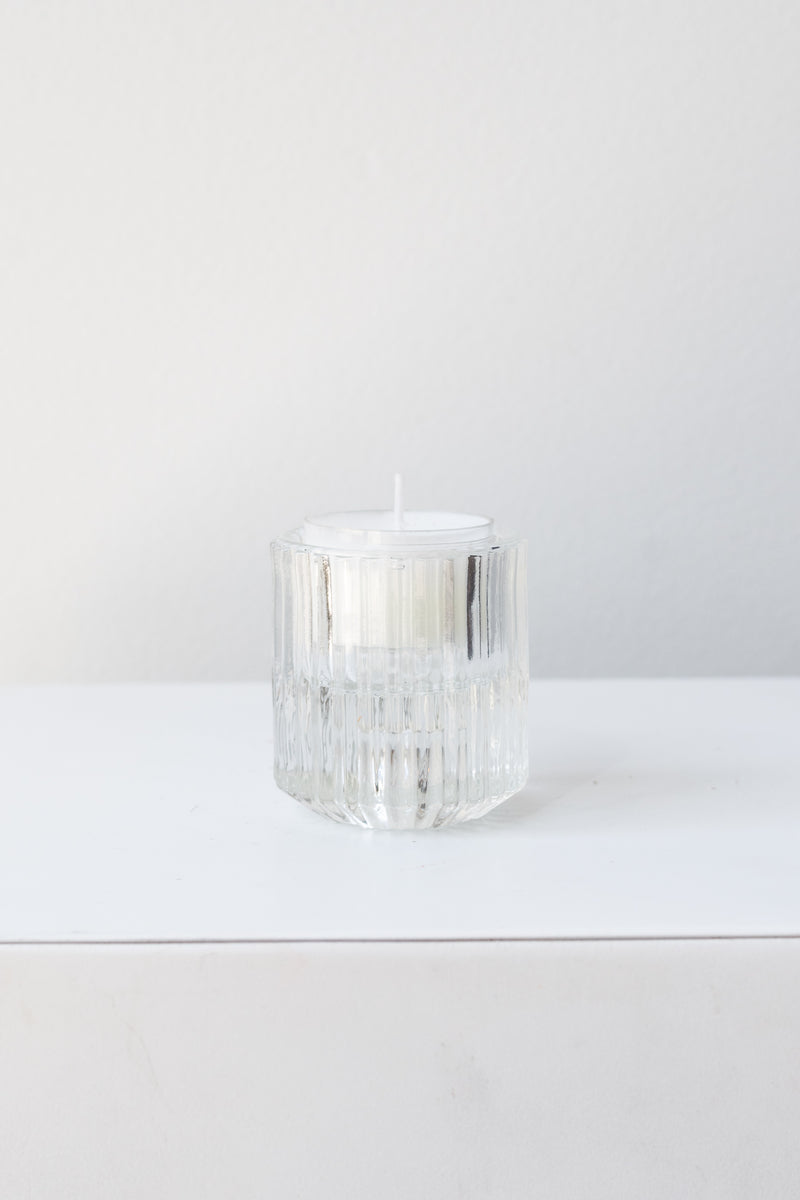 White tea light candle inside clear pleated glass candle holder in front of white background