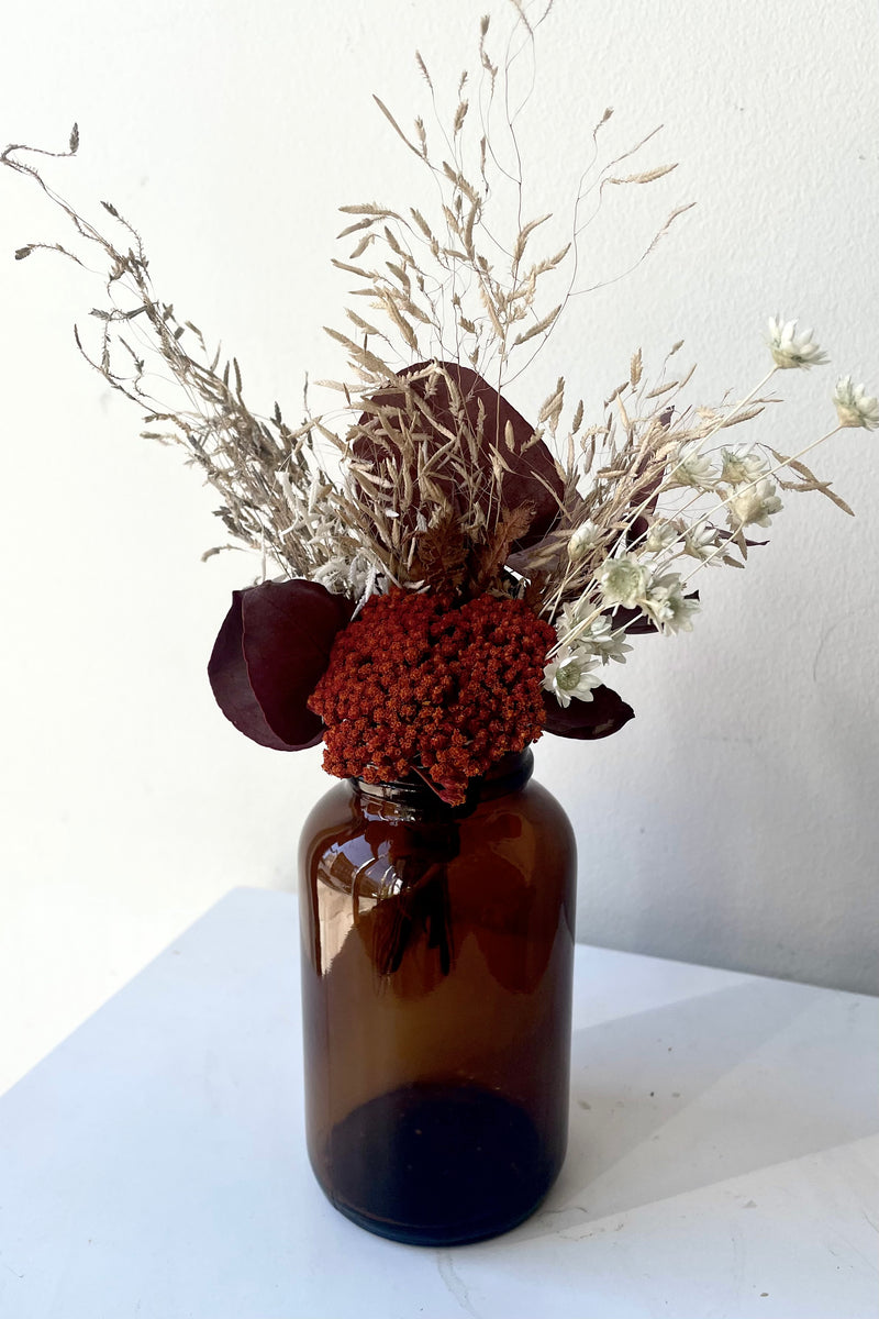 A dried floral bouquet arrangement made by Sprout Home sits in the 250cc Amber Glass Round Bottle against a white backdrop