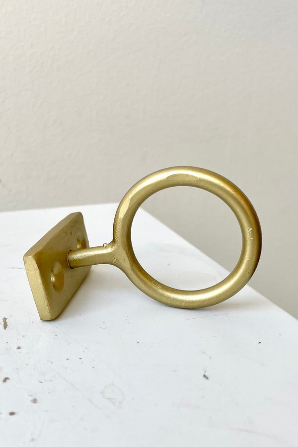 The brass Bijou hanging hardware against. white wall showing the mount and attached ring at Sprout Home.