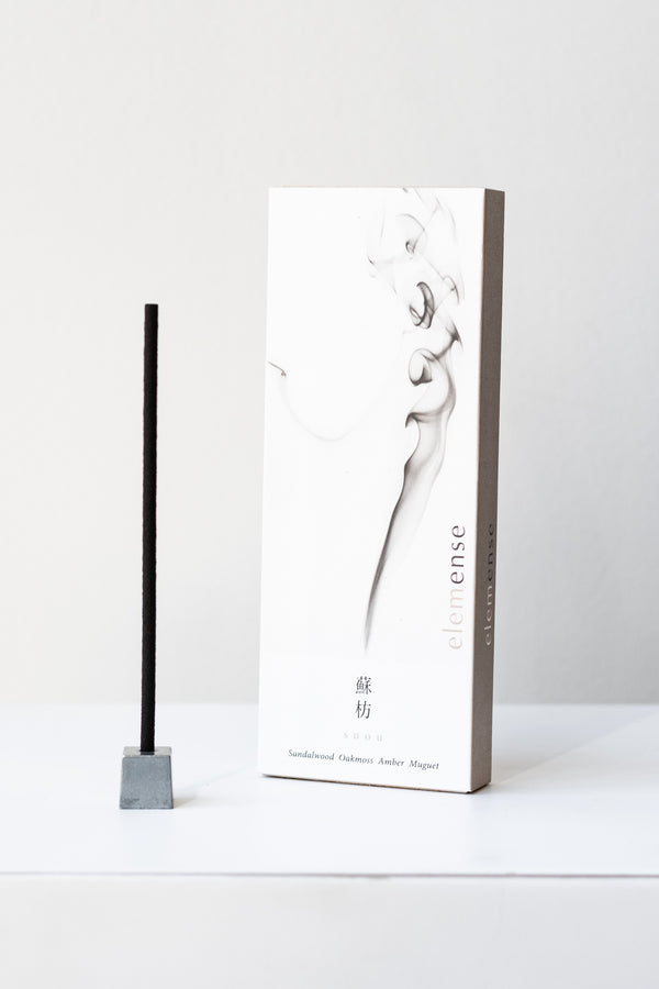 A white and grey rectangular box of Elemense incense sits on a white table in a white room. To the left of the box is a small silver cube incense holder with an unlit stick of black incense in it. This photo is of the Suou incense.