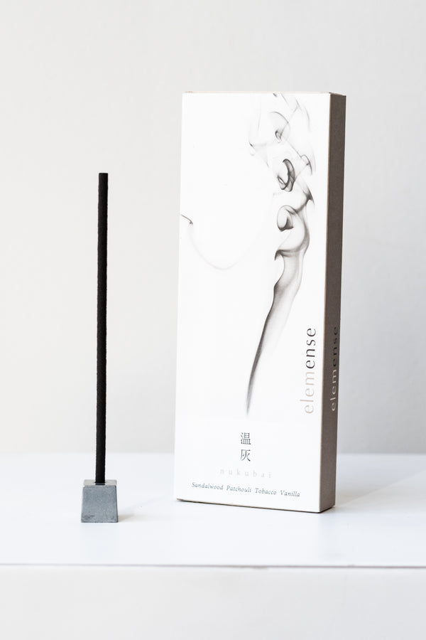 A white and grey rectangular box of Elemense incense sits on a white table in a white room. To the left of the box is a small silver cube incense holder with an unlit stick of black incense in it. This photo is of the Nukubai incense.