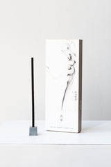 A white and grey rectangular box of Elemense incense sits on a white table in a white room. To the left of the box is a small silver cube incense holder with an unlit stick of black incense in it. This photo is of the Tetsukon incense.