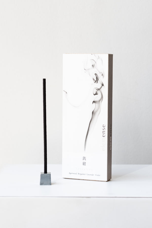 A white and grey rectangular box of Elemense incense sits on a white table in a white room. To the left of the box is a small silver cube incense holder with an unlit stick of black incense in it. This photo is of the Tetsukon incense.