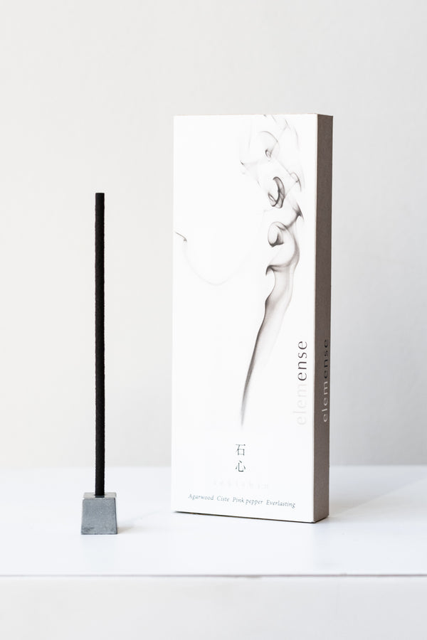 A white and grey rectangular box of Elemense incense sits on a white table in a white room. To the left of the box is a small silver cube incense holder with an unlit stick of black incense in it. This photo is of the Sekishin incense.