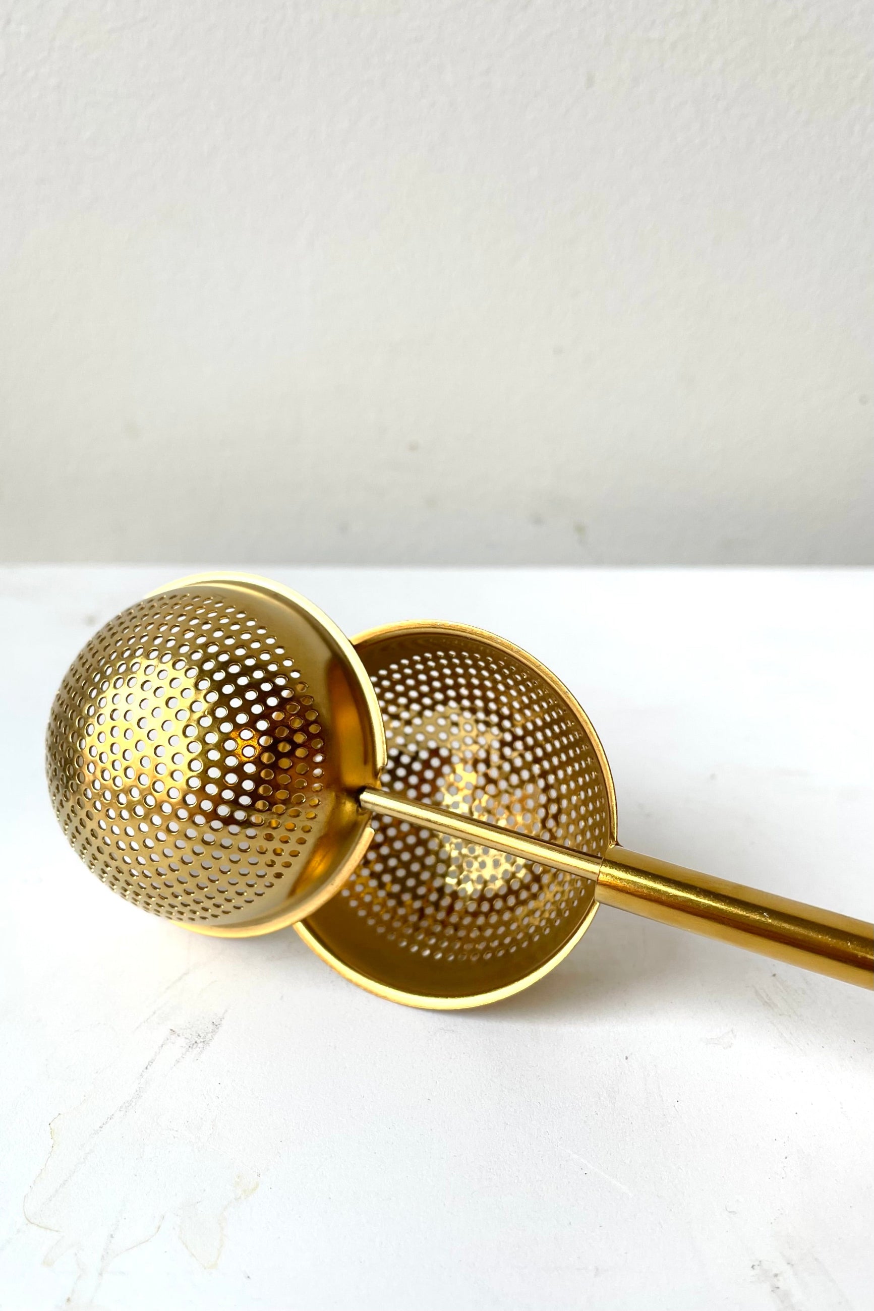 A stainless steel tea egg with a perforated design and a gold finish. The tea egg is shown disassembled, with its spherical upper part separated from the elongated handle. ©Sprout Home