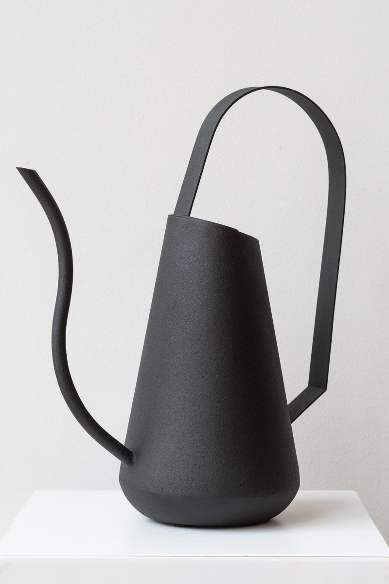 Black Metal Tall Watering Can on white pedestal in front of white background