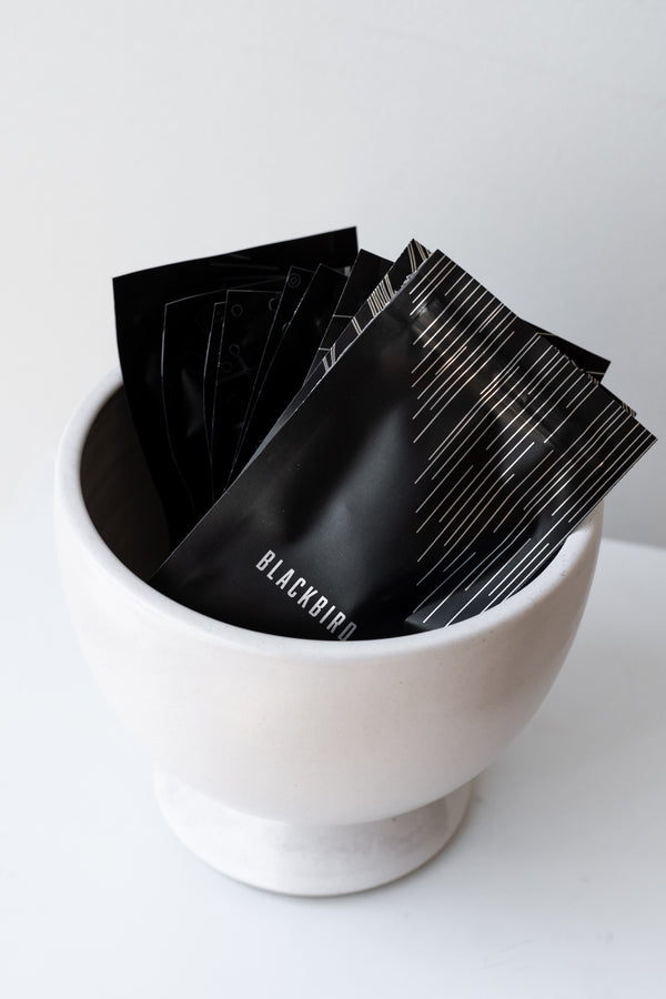 Packets of incense cones from the Blackbird Incense Discovery set sit in a white bowl on a white surface in a white room. Each packet is black plastic with a silver geometric design.
