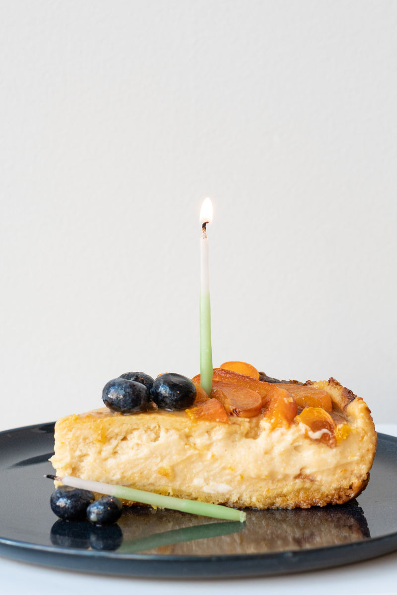 Knot and Bow beeswax candle stuck in slice of cheesecake on blue plate with blueberries and apricots in front of white background