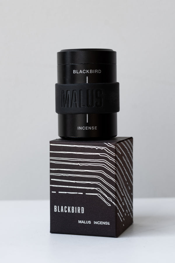A black box of Blackbird incense sits on a white surface in a white room. This incense is called MALUS and has a white line design on the box. On top of the box sits a black cylindrical container that holds the incense cones. The container has a black rubber band around the middle with "Malus" etched in it.