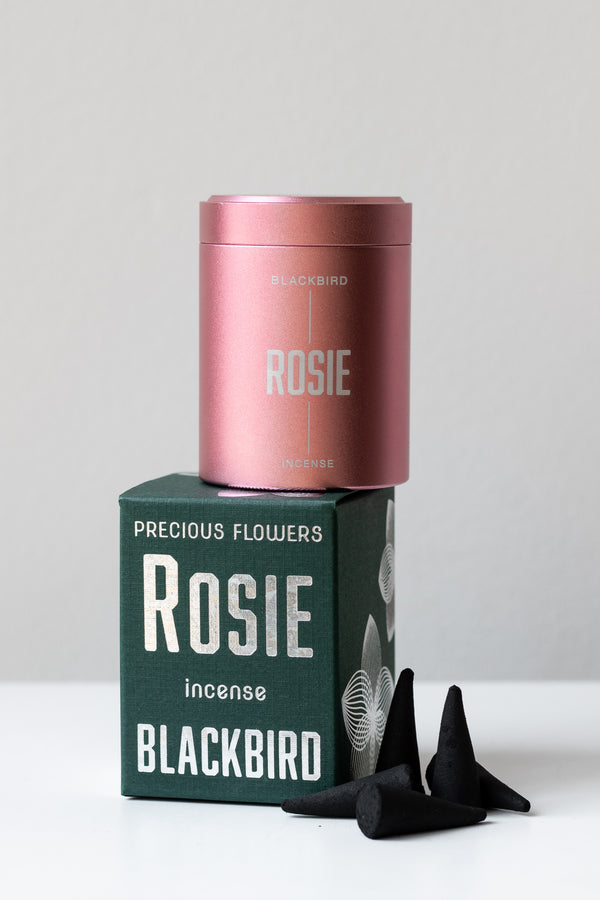 Rosie incense by BLACKBIRD sits on a white surface in a white room. The pink incense tin is on top of the box, and five black incense cones are scattered to the side of the box. 