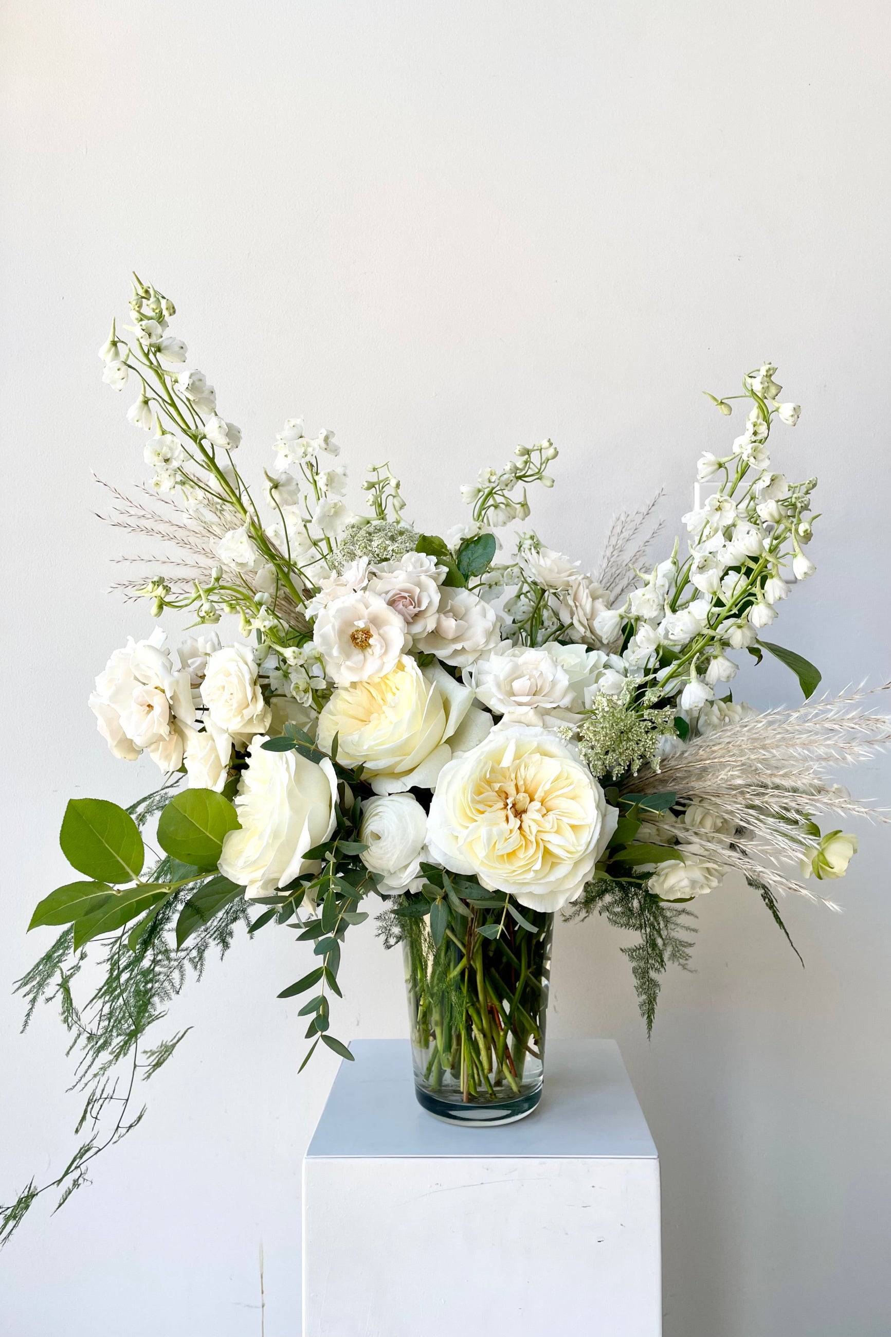 A neutral white elegant arrangement with white delphinium, garden roses and pampas plooms by Sprout Home against a white wall. ©Sprout Home #size_$160