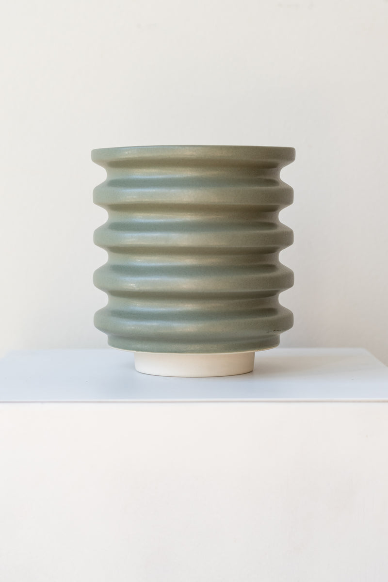 A grey-green planter sits on a white surface in a white room. The planter is cylindrical and ribbed. It sits on an unglazed ceramic drainage tray. The planter is empty. It is photographed straight on.