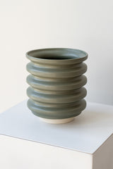 A grey-green planter sits on a white surface in a white room. The planter is cylindrical and ribbed. It sits on an unglazed ceramic drainage tray. The planter is empty. It is photographed closer and at an angle.