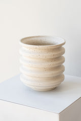 A white planter with black speckles sits on a white surface in a white room. The planter is cylindrical and ribbed. It sits on an unglazed ceramic drainage tray. The planter is empty. It is photographed closer and at an angle.