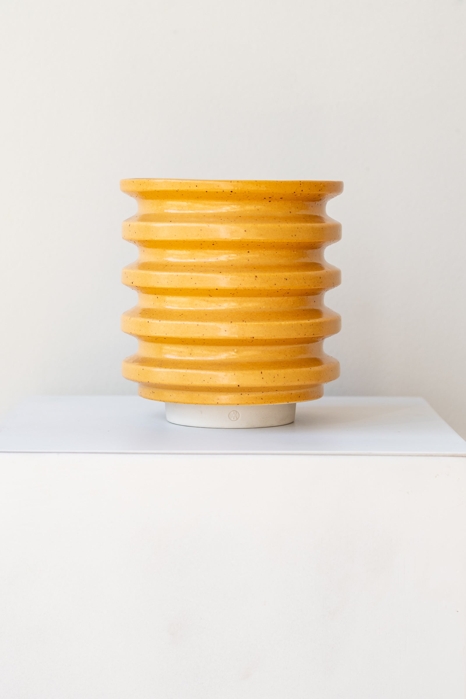 A yellow planter with black speckles sits on a white surface in a white room. The planter is cylindrical and ribbed. It sits on an unglazed ceramic drainage tray. The planter is empty. It is photographed straight on.