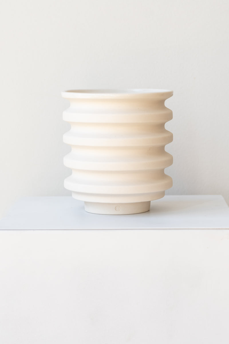 A white planter sits on a white surface in a white room. The planter is cylindrical and ribbed. It sits on an unglazed ceramic drainage tray. The planter is empty. It is photographed straight on.
