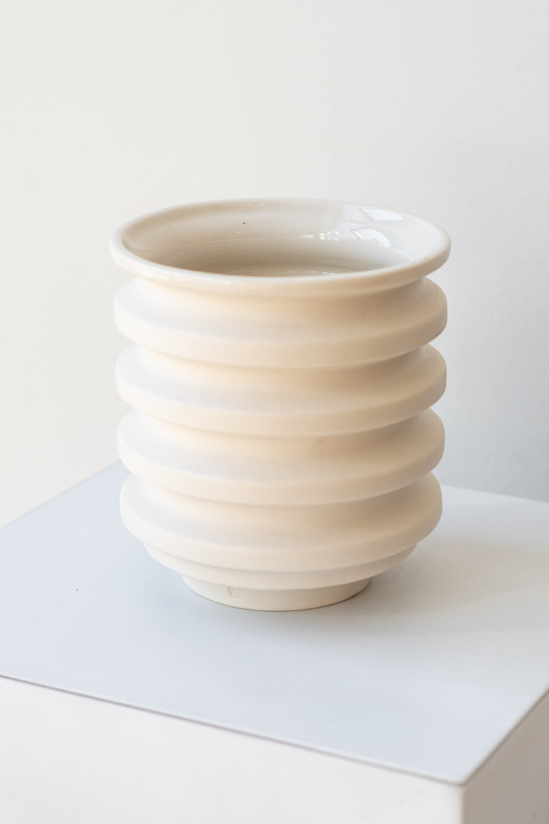 A white planter sits on a white surface in a white room. The planter is cylindrical and ribbed. It sits on an unglazed ceramic drainage tray. The planter is empty. It is photographed closer and at an angle.
