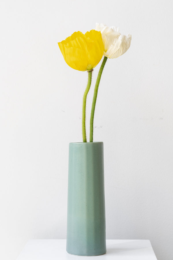Bloom Vase in rosemary by The Bright Angle sits on a white surface in a white room. Inside the vase are two poppies, one yellow and one white. 