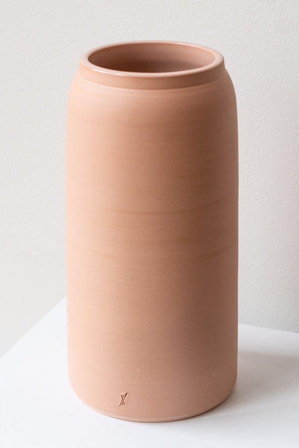 One large pink ceramic bouquet vase sits on a white surface in a white room. The vase is empty. It is photographed closer and at an angle.