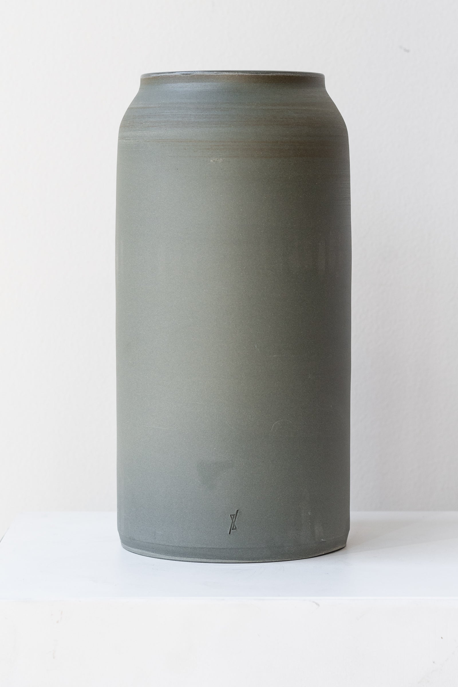 One large green-grey ceramic bouquet vase sits on a white surface in a white room. The vase is empty. It is photographed straight on.