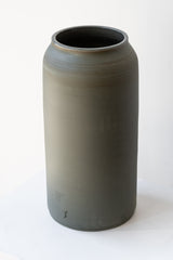 One large green-grey ceramic bouquet vase sits on a white surface in a white room. The vase is empty. It is photographed closer and at an angle.