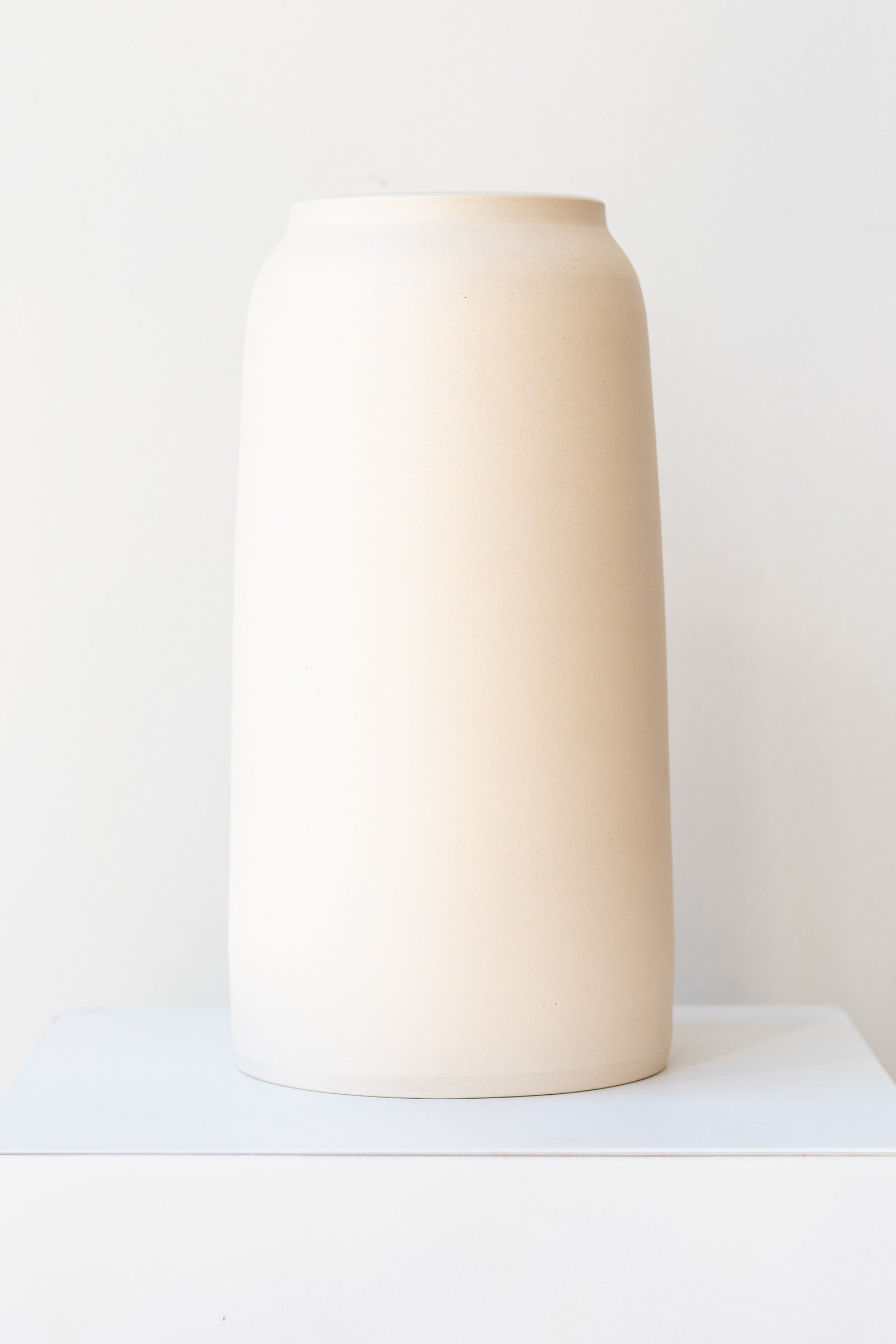 One cream colored bouquet vase sits on a white surface in a white room. The vase is tall and cylindrical, with a slight rim at the top. It is photographed straight on.