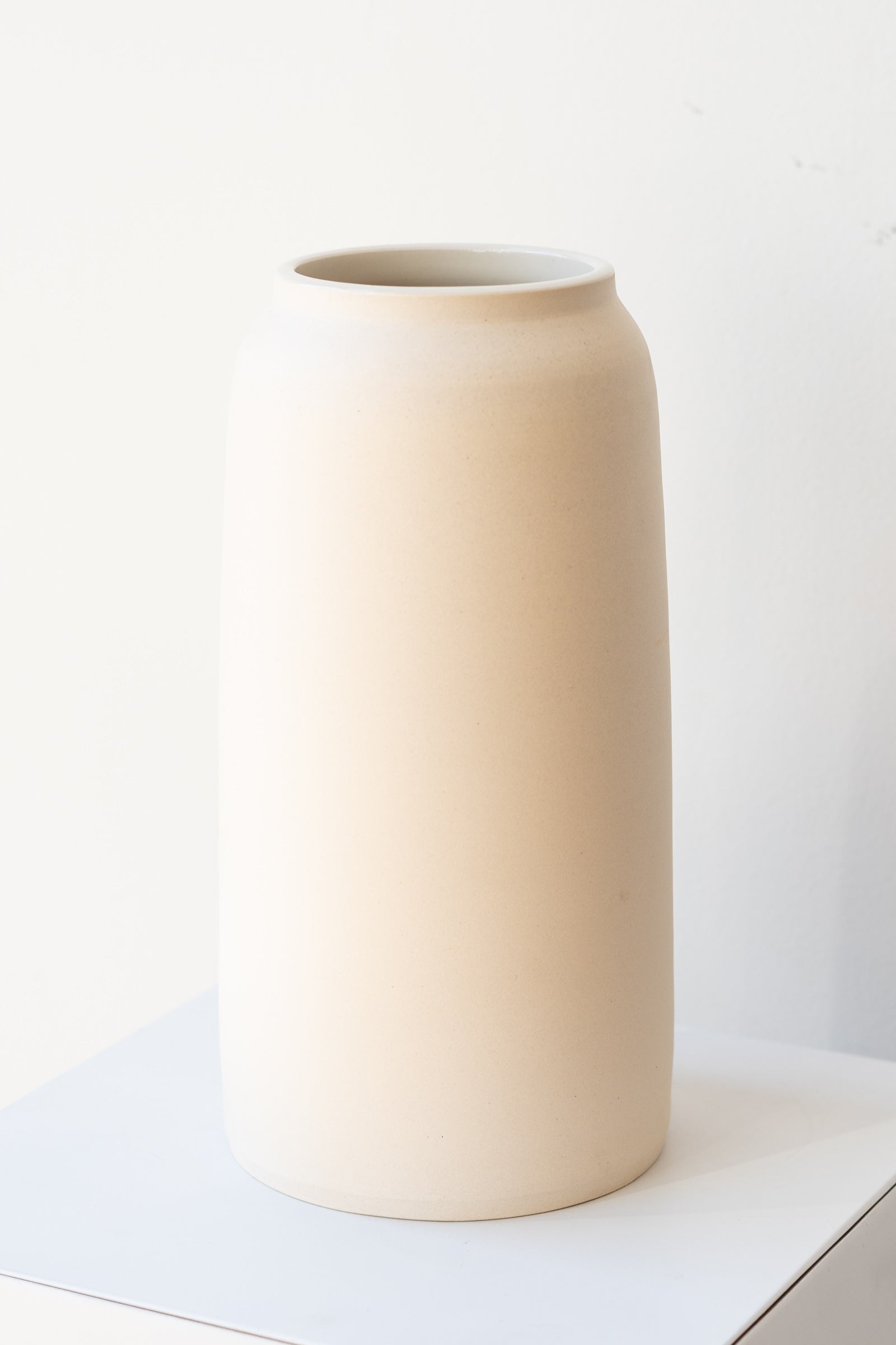 One cream colored bouquet vase sits on a white surface in a white room. The vase is tall and cylindrical, with a slight rim at the top. It is photographed at a slight angle to show the clay detail.