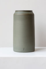 One medium green-grey stoneware vase sits on a white surface in a white room. It is round and tall, and it has a small logo imprinted at the base of the vase. It is photographed straight on.