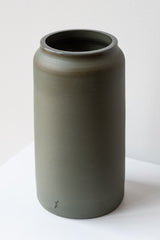 One medium green-grey stoneware vase sits on a white surface in a white room. It is round and tall, and it has a small logo imprinted at the base of the vase. It is photographed closer and at an angle.