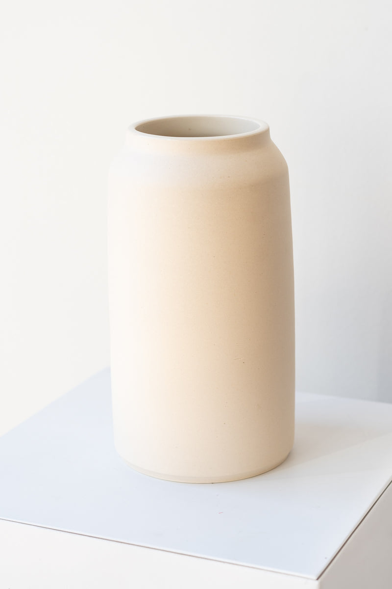 One cream colored bouquet vase sits on a white surface in a white room. The vase is tall and cylindrical, with a slight rim at the top. It is photographed at a slight angle to show the clay detail.