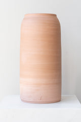 One extra large pink ceramic bouquet vase sits on a white surface in a white room. The vase is empty. It is photographed straight on.