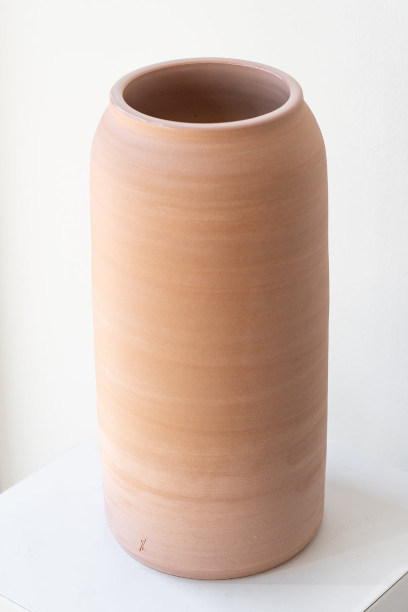 One extra large pink ceramic bouquet vase sits on a white surface in a white room. The vase is empty. It is photographed closer and at an angle.
